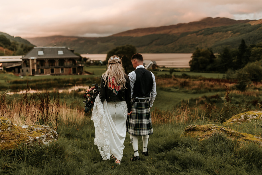 Bride wearing leather jacket and groom wearing kilt walk through grass